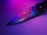 A close-up view of a knife with a metallic blade featuring a subtle spider logo. The blade reflects vibrant neon colors, creating a gradient effect of purples and blues. The background is softly illuminated, enhancing the colors on the knife.