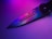 A close-up view of a knife with a metallic blade featuring a subtle spider logo. The blade reflects vibrant neon colors, creating a gradient effect of purples and blues. The background is softly illuminated, enhancing the colors on the knife.