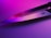 A sharp knife blade reflecting pink and purple lighting, creating a vibrant and modern aesthetic. The metal surface shows a logo engraved near the handle.