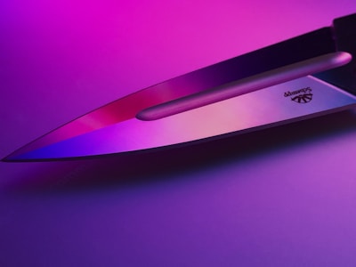 A sharp knife blade reflecting pink and purple lighting, creating a vibrant and modern aesthetic. The metal surface shows a logo engraved near the handle.