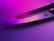 A sharp knife blade reflecting pink and purple lighting, creating a vibrant and modern aesthetic. The metal surface shows a logo engraved near the handle.