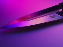 A sharp knife blade reflecting pink and purple lighting, creating a vibrant and modern aesthetic. The metal surface shows a logo engraved near the handle.