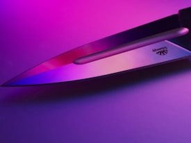 A sharp knife blade reflecting pink and purple lighting, creating a vibrant and modern aesthetic. The metal surface shows a logo engraved near the handle.