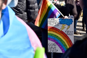 A person holds a rainbow flag and a colorful protest sign that reads 'Some parents are their child's first bully... GSA's save lives!' The background shows other people, some wearing jackets.