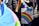 A person holds a rainbow flag and a colorful protest sign that reads 'Some parents are their child's first bully... GSA's save lives!' The background shows other people, some wearing jackets.