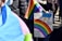A person holds a rainbow flag and a colorful protest sign that reads 'Some parents are their child's first bully... GSA's save lives!' The background shows other people, some wearing jackets.
