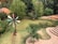 A small pond is surrounded by lush green vegetation and two colorful windmills. Steps made of brick lead down to a landscaped area near the pond. Various shades of green foliage provide a backdrop, creating a serene and natural setting.