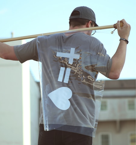 A person wearing a gray cap and T-shirt holds a transparent flag over their shoulder. The flag features a white design with symbols and a heart. The scene is outdoors with a building in the background.