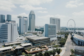 A cityscape featuring modern skyscrapers and high-rise buildings, including notable hotels and landmarks. A large ferris wheel is visible in the background near a body of water, and there are roads lined with trees. The sky is partly cloudy, creating a bright yet relaxed atmosphere.