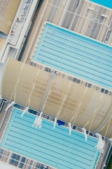 An aerial view of two large swimming pools, one covered partially by a curved roof structure. The pools are surrounded by a deck with a striped pattern. Lanes are visible in the pools, indicating their use for swimming.