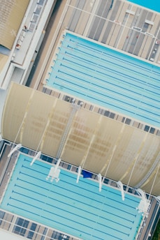 An aerial view of two large swimming pools, one covered partially by a curved roof structure. The pools are surrounded by a deck with a striped pattern. Lanes are visible in the pools, indicating their use for swimming.