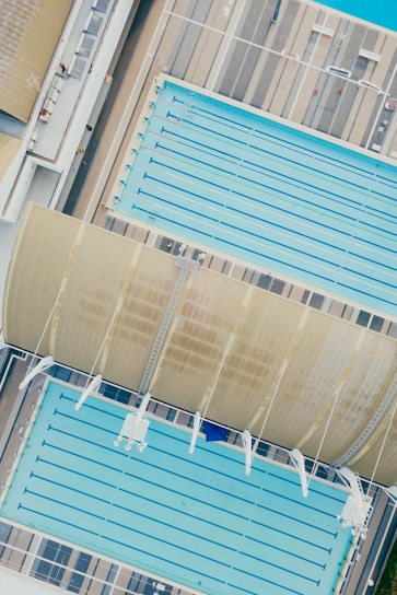 An aerial view of two large swimming pools, one covered partially by a curved roof structure. The pools are surrounded by a deck with a striped pattern. Lanes are visible in the pools, indicating their use for swimming.