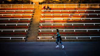 A person is roller skating on a concrete surface in front of a large, empty tiered seating area. The seating area is comprised of long rows of benches painted red and white, with a central set of stairs leading upwards to the top. Two individuals are sitting at the top of the stairs, engaged in conversation.