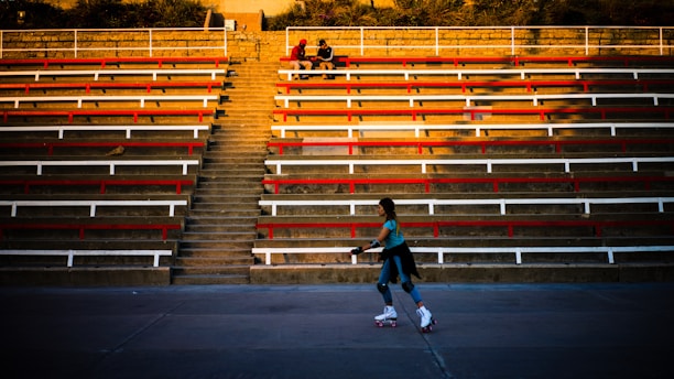 A person is roller skating on a concrete surface in front of a large, empty tiered seating area. The seating area is comprised of long rows of benches painted red and white, with a central set of stairs leading upwards to the top. Two individuals are sitting at the top of the stairs, engaged in conversation.