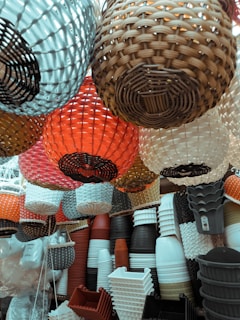 A collection of variously colored and styled baskets and planters are displayed in a market setting. The baskets are hanging from above, showcasing their intricate woven designs. Planters in different shapes and colors are stacked below, creating a layered effect.