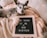 A small dog is lying comfortably on beige blankets, next to a letter board. The board displays the message, 'I'M GOING TO BE A BIG SISTER,' indicating a playful or affectionate announcement likely related to a family expecting a child.