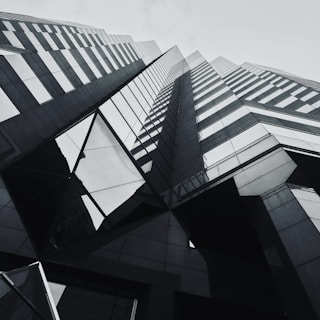 The architectural image features an upward view of a modern skyscraper, characterized by sleek lines and reflective glass surfaces. The building's design includes geometric patterns and sharp angles, creating a sense of grandeur and innovation.