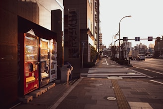 A city street scene features a row of vending machines brightly lit by the setting sun. The machines contain various beverages and are flanked by a reflective building facade on the left. The street is relatively empty with a few vehicles in the distance, and there are crossing lines and sidewalks visible.