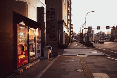 A city street scene features a row of vending machines brightly lit by the setting sun. The machines contain various beverages and are flanked by a reflective building facade on the left. The street is relatively empty with a few vehicles in the distance, and there are crossing lines and sidewalks visible.