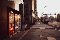 A city street scene features a row of vending machines brightly lit by the setting sun. The machines contain various beverages and are flanked by a reflective building facade on the left. The street is relatively empty with a few vehicles in the distance, and there are crossing lines and sidewalks visible.