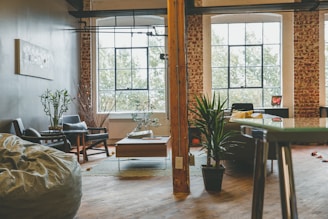 A stylish and cozy living space featuring a mix of modern and rustic elements. Large windows with a grid pattern provide natural light, highlighting the exposed brick walls and wooden floor. Potted plants add a touch of greenery, while contemporary furniture includes a bean bag, chairs, and a sofa with colorful cushions. A coffee table is centered on a small area rug, and the room is divided by a wooden beam.