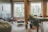 A stylish and cozy living space featuring a mix of modern and rustic elements. Large windows with a grid pattern provide natural light, highlighting the exposed brick walls and wooden floor. Potted plants add a touch of greenery, while contemporary furniture includes a bean bag, chairs, and a sofa with colorful cushions. A coffee table is centered on a small area rug, and the room is divided by a wooden beam.
