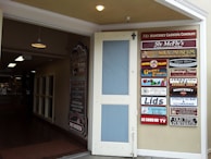 An open door leading to an indoor space with various lighting fixtures hanging from the ceiling. On the right, a wall displays multiple colorful signs for different businesses and attractions, including a wax museum, gourmet express, and vintage photography.