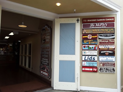 An open door leading to an indoor space with various lighting fixtures hanging from the ceiling. On the right, a wall displays multiple colorful signs for different businesses and attractions, including a wax museum, gourmet express, and vintage photography.