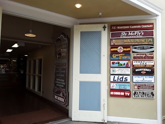 An open door leading to an indoor space with various lighting fixtures hanging from the ceiling. On the right, a wall displays multiple colorful signs for different businesses and attractions, including a wax museum, gourmet express, and vintage photography.