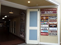 An open door leading to an indoor space with various lighting fixtures hanging from the ceiling. On the right, a wall displays multiple colorful signs for different businesses and attractions, including a wax museum, gourmet express, and vintage photography.