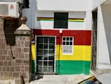 A small building facade is painted with horizontal red, yellow, and green stripes. There is a black metal gate at the entrance and a window adjacent to the door. An air conditioning unit is mounted on the brick wall next to the building, and a large stone pillar is visible to the left.