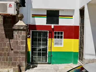 A small building facade is painted with horizontal red, yellow, and green stripes. There is a black metal gate at the entrance and a window adjacent to the door. An air conditioning unit is mounted on the brick wall next to the building, and a large stone pillar is visible to the left.