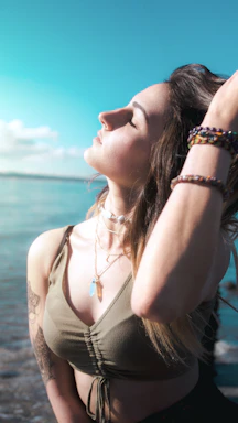 A woman wearing layered gold bracelets and earrings, standing against a soft ocean backdrop.