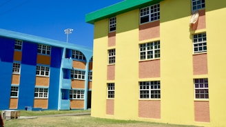 Colorful apartment buildings featuring bright blue and yellow walls with contrasting orange panels. The structures have multiple windows, and there is a cloth hanging from one of the windows. The area around the buildings has a clear sky and an open grassy space.