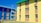 Colorful apartment buildings featuring bright blue and yellow walls with contrasting orange panels. The structures have multiple windows, and there is a cloth hanging from one of the windows. The area around the buildings has a clear sky and an open grassy space.