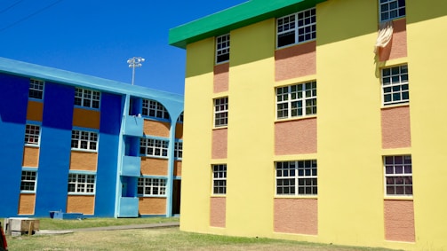 Colorful apartment buildings featuring bright blue and yellow walls with contrasting orange panels. The structures have multiple windows, and there is a cloth hanging from one of the windows. The area around the buildings has a clear sky and an open grassy space.