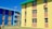 Colorful apartment buildings featuring bright blue and yellow walls with contrasting orange panels. The structures have multiple windows, and there is a cloth hanging from one of the windows. The area around the buildings has a clear sky and an open grassy space.