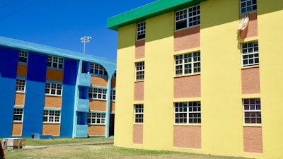 Colorful apartment buildings featuring bright blue and yellow walls with contrasting orange panels. The structures have multiple windows, and there is a cloth hanging from one of the windows. The area around the buildings has a clear sky and an open grassy space.