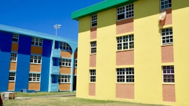 Colorful apartment buildings featuring bright blue and yellow walls with contrasting orange panels. The structures have multiple windows, and there is a cloth hanging from one of the windows. The area around the buildings has a clear sky and an open grassy space.
