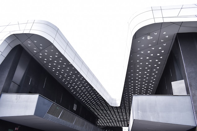 Modern architectural structure featuring sleek lines and a geometric roof pattern with triangular cutouts. The building exterior has a predominantly metallic and dark color scheme, highlighted by a reflective surface.
