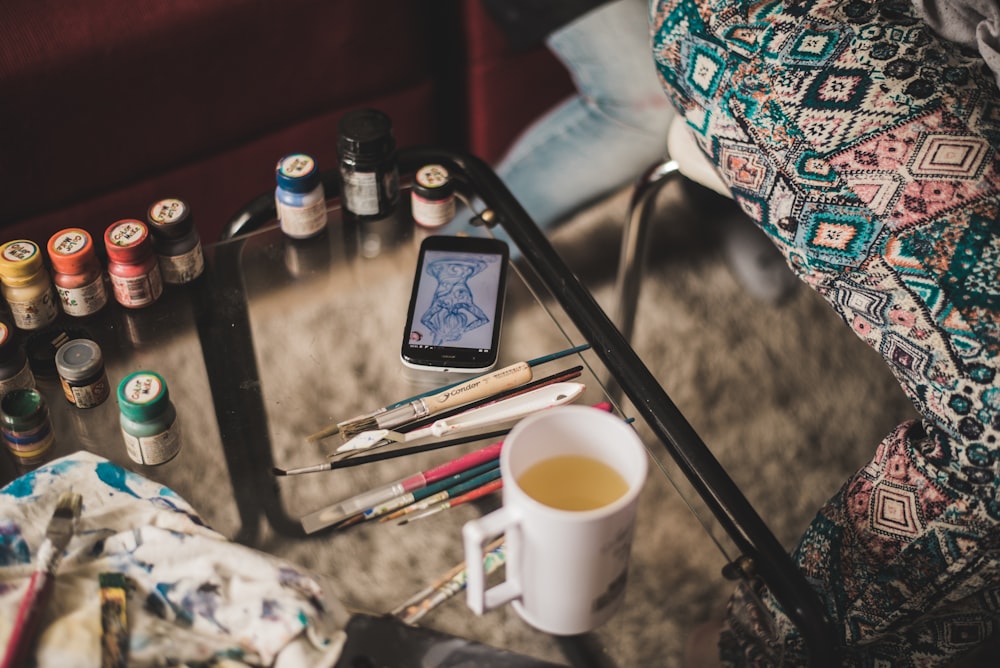 white mug with yellow beverage beside paint brushed on clear glass table