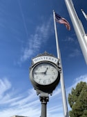 An ornate street clock with the word 'Monterey' displayed at the top, set against a clear blue sky with wispy clouds. The clock shows the time as approximately 1:54. To the right, a tall flagpole holds an American flag that is waving gently in the breeze.