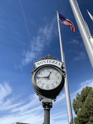 An ornate street clock with the word 'Monterey' displayed at the top, set against a clear blue sky with wispy clouds. The clock shows the time as approximately 1:54. To the right, a tall flagpole holds an American flag that is waving gently in the breeze.