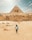 A person walks across a desert landscape with the Great Pyramid and the Sphinx of Giza in the background under a partially cloudy sky. The sand-colored stones of the ancient structures contrast with the vast expanse of sand.