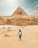 A person walks across a desert landscape with the Great Pyramid and the Sphinx of Giza in the background under a partially cloudy sky. The sand-colored stones of the ancient structures contrast with the vast expanse of sand.