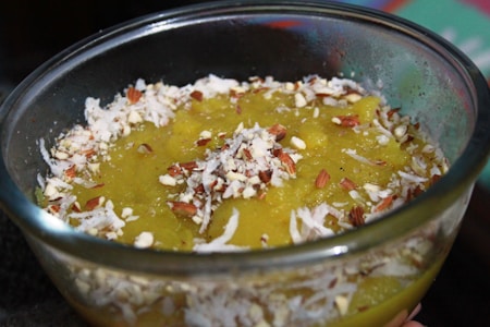 A glass bowl filled with a yellow pudding-like dessert, topped with shredded coconut and chopped nuts, is in focus. The dessert has a rich and creamy texture with visible coconut flakes and pieces of almonds scattered on top, enhancing its appearance.