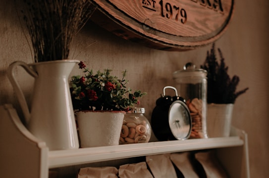 A rustic kitchen shelf adorned with a variety of decorative items including a white ceramic pitcher holding dried flowers, a small potted plant with red berries, and a jar filled with cookies or pastries. A vintage-style black alarm clock is placed beside a glass jar filled with rings of dried fruit. The overall setting is enhanced by a circular wooden sign on the wall above the shelf, adding to the rustic ambiance.