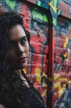 A person with long, dark curly hair and red lipstick stands in front of a colorful graffiti wall. The vivid street art on the wall features splashes of red, green, and yellow, creating a lively and urban backdrop.