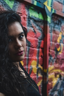 A person with long, dark curly hair and red lipstick stands in front of a colorful graffiti wall. The vivid street art on the wall features splashes of red, green, and yellow, creating a lively and urban backdrop.