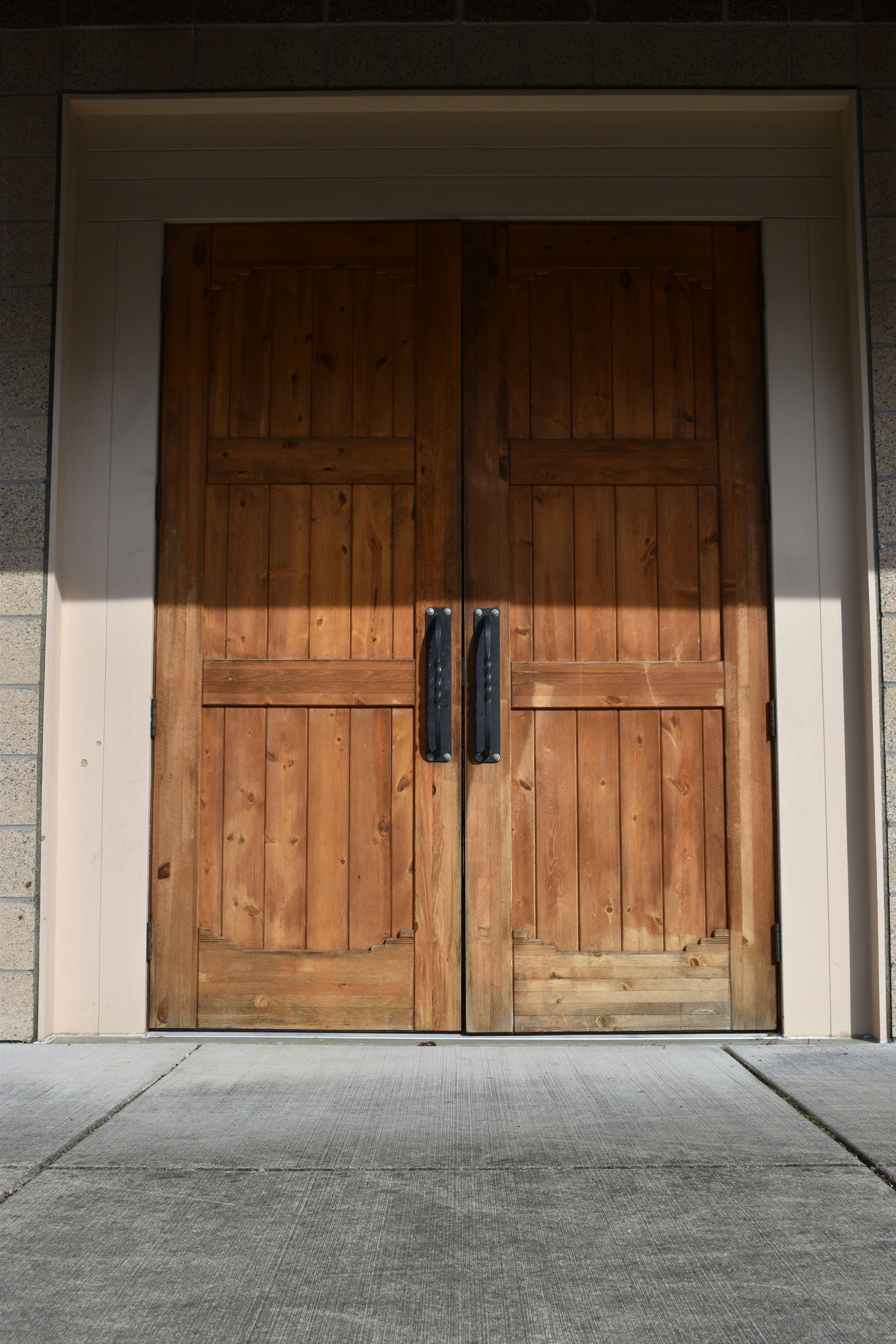 A pair of large wooden double doors with vertical panels and two dark metal handles. The doors are set in a light-colored frame, and the surrounding area includes textured brick walls and a concrete floor.